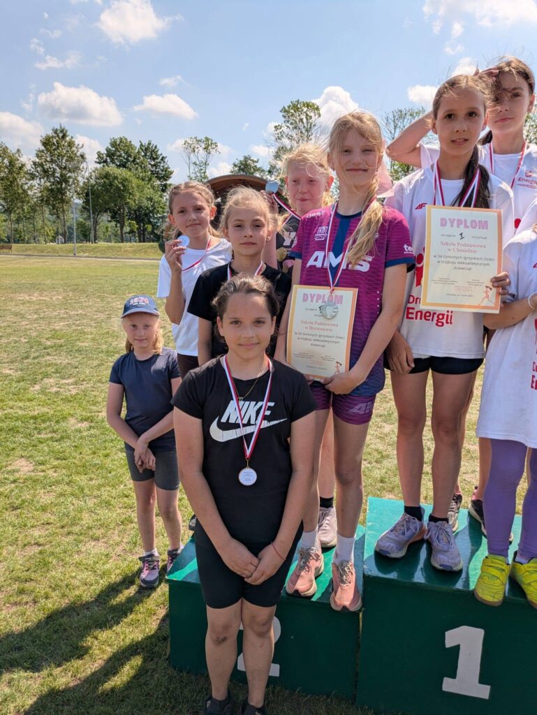 Dziewczęta ze szkoły podstawowej stoją na podium po zawodach lekkoatletycznych, prezentując medale i dyplomy zdobyte w rywalizacji drużynowej.
