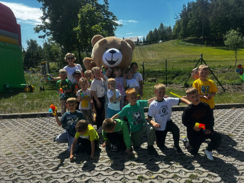 Dzieci z oddziału przedszkolnego wraz z opiekunką pozują do zdjęcia z dużą maskotką misia podczas festynu i zabaw na świeżym powietrzu.