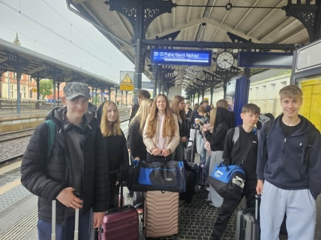 Grupa uczniów z walizkami czeka na peronie dworca kolejowego na odjazd pociągu. Młodzież przygotowuje się do wyjazdu edukacyjnego, a w tle widoczne są tory, zegar i tablica z informacją o odjeździe.