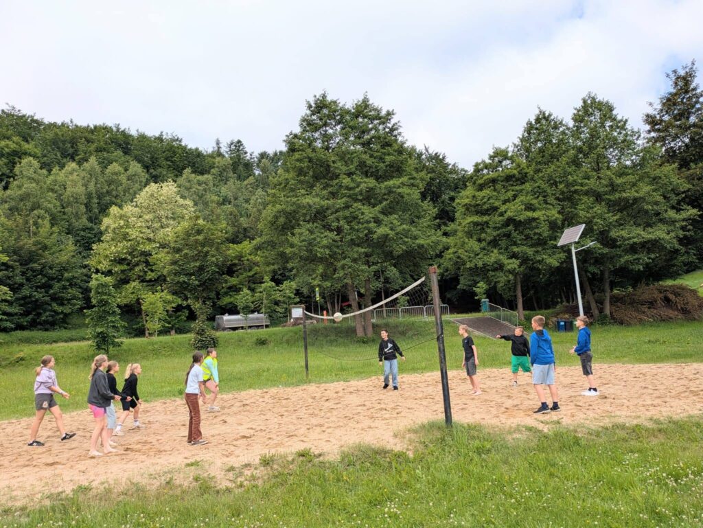Grupa uczniów gra w siatkówkę plażową na piaszczystym boisku położonym wśród zieleni. Wspólna aktywność na świeżym powietrzu sprzyja integracji i dobrej zabawie podczas wyjazdu.