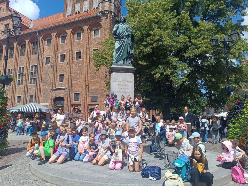 Grupa uczniów wraz z opiekunami pozuje do pamiątkowego zdjęcia przy pomniku Mikołaja Kopernika. W tle widoczna jest zabytkowa ceglana zabudowa toruńskiej starówki oraz słoneczna letnia pogoda.