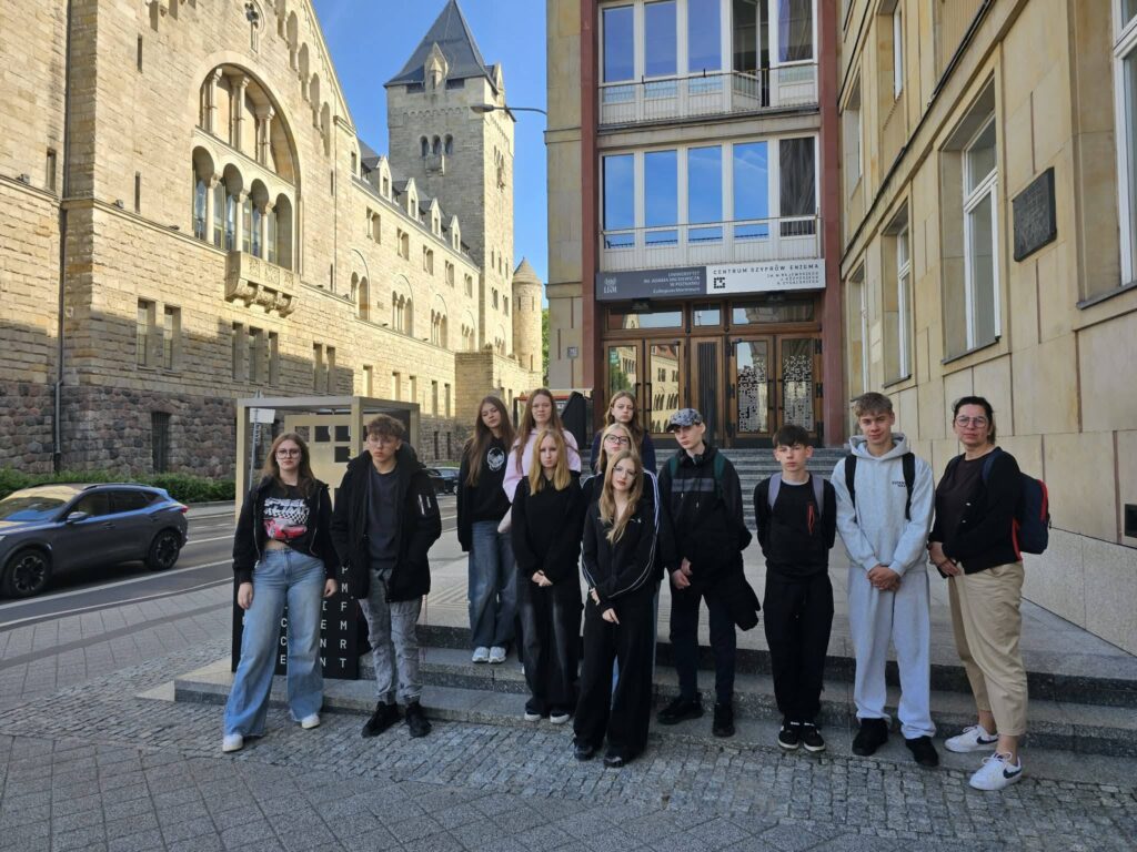 Uczniowie wraz z opiekunką pozują do wspólnego zdjęcia przed wejściem do Centrum Szyfrów Enigma. W tle widoczne są zabytkowe budynki oraz miejska zabudowa.