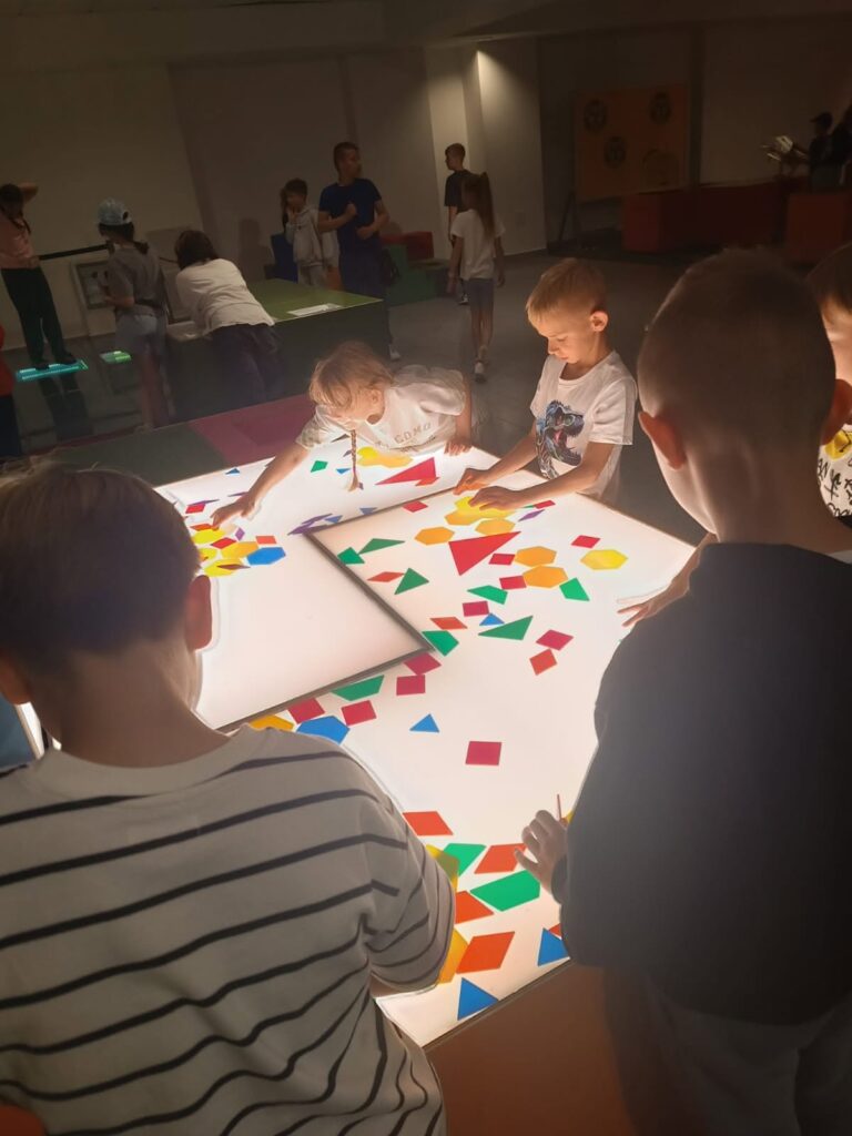 Dzieci wspólnie bawią się i uczą przy podświetlanym stole edukacyjnym. Układają kolorowe figury geometryczne, rozwijając wyobraźnię przestrzenną, logiczne myślenie i współpracę.