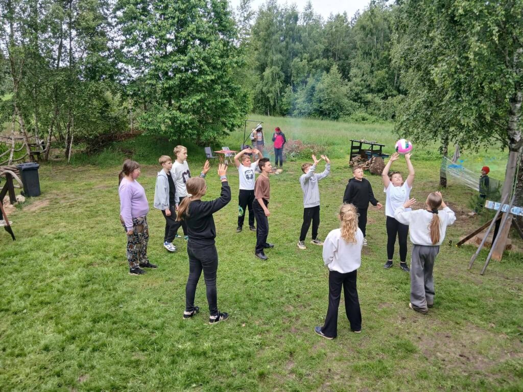 Grupa uczni贸w sp臋dza czas na 艣wie偶ym powietrzu, wsp贸lnie graj膮c w pi艂k臋 na trawiastej polanie otoczonej drzewami. W tle widoczne jest miejsce na ognisko, a dzieci aktywnie uczestnicz膮 w zabawie.