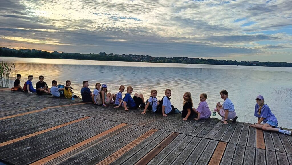 Grupa dzieci siedzi na drewnianym pomoście nad jeziorem, odpoczywając i podziwiając spokojną taflę wody. W tle widoczne są chmury na niebie oraz przeciwległy brzeg jeziora.