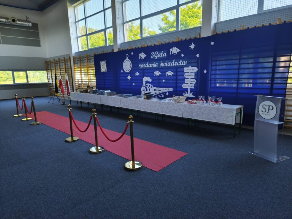 Sala gimnastyczna przygotowana do uroczystej Gali Rozdania Świadectw. Widoczna jest okolicznościowa dekoracja, czerwony dywan, stół z nagrodami i świadectwami oraz miejsce przeznaczone do wręczania wyróżnień uczestnikom uroczystości.