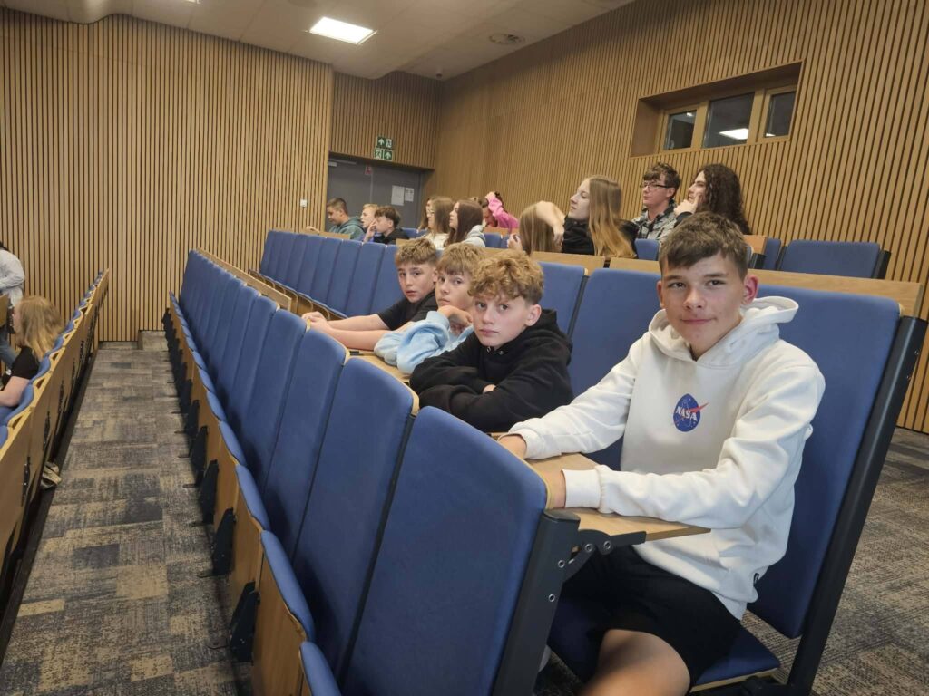 Uczniowie siedzą w sali wykładowej i uczestniczą w zajęciach lub prezentacji edukacyjnej.