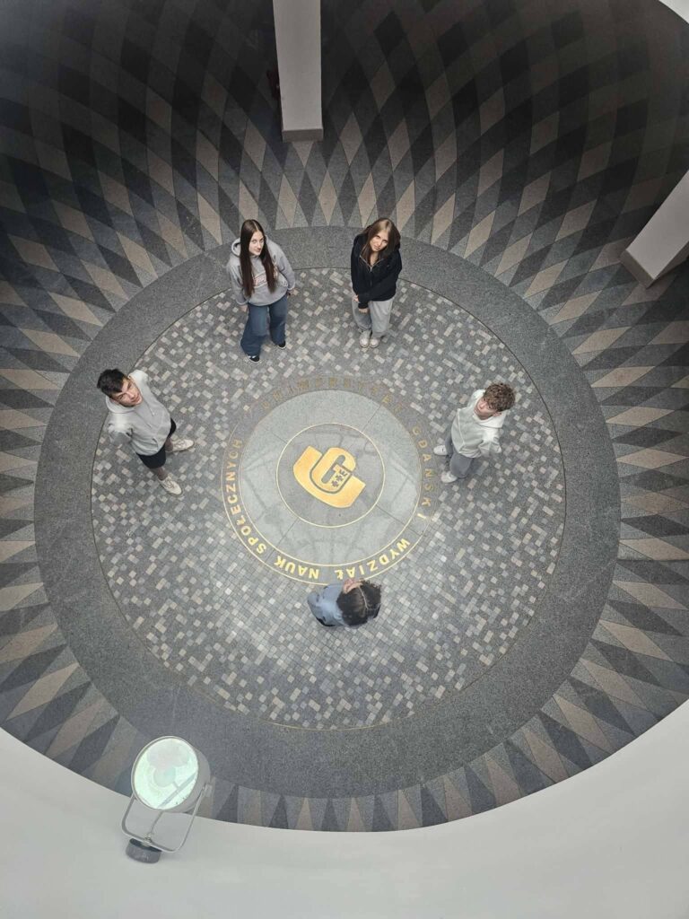 Uczniowie stoją wokół okrągłej mozaiki z logo Uniwersytetu Gdańskiego w holu budynku.