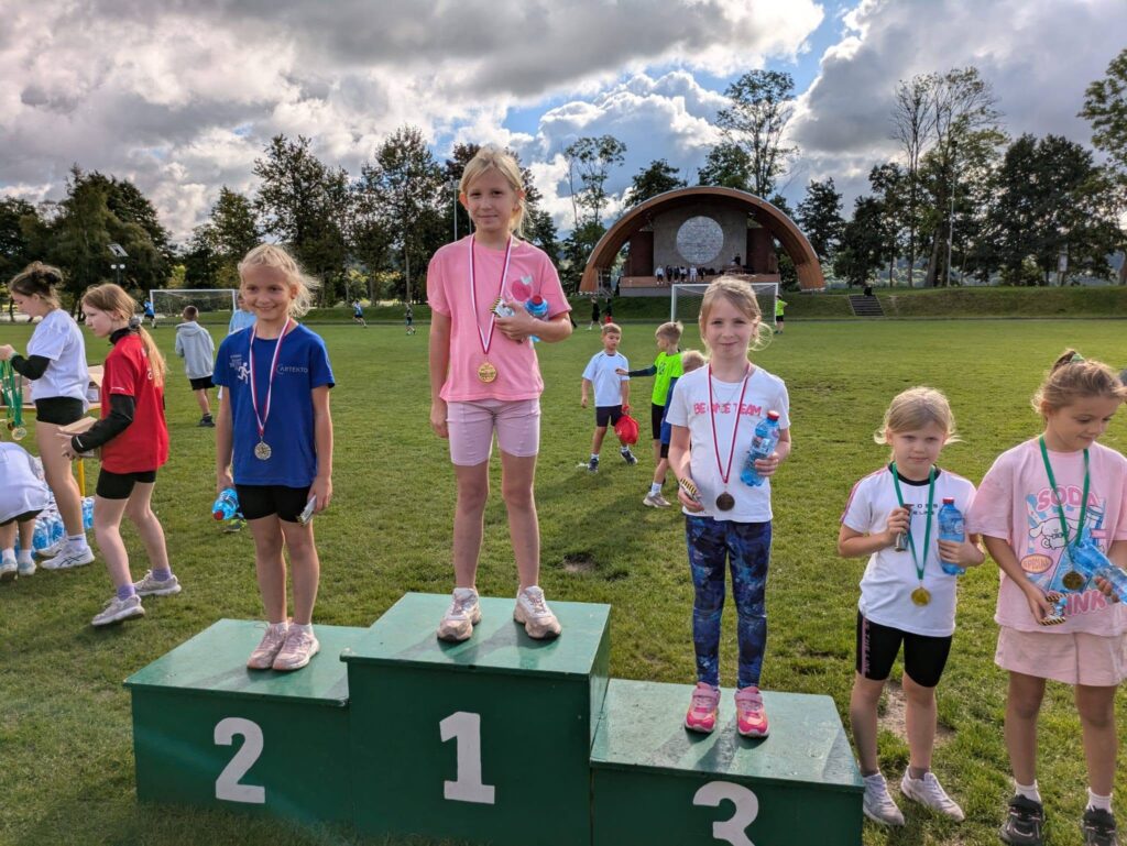 Uczennice stoją na podium podczas zawodów sportowych na boisku. Dziewczynki mają na szyjach medale i trzymają butelki z wodą.